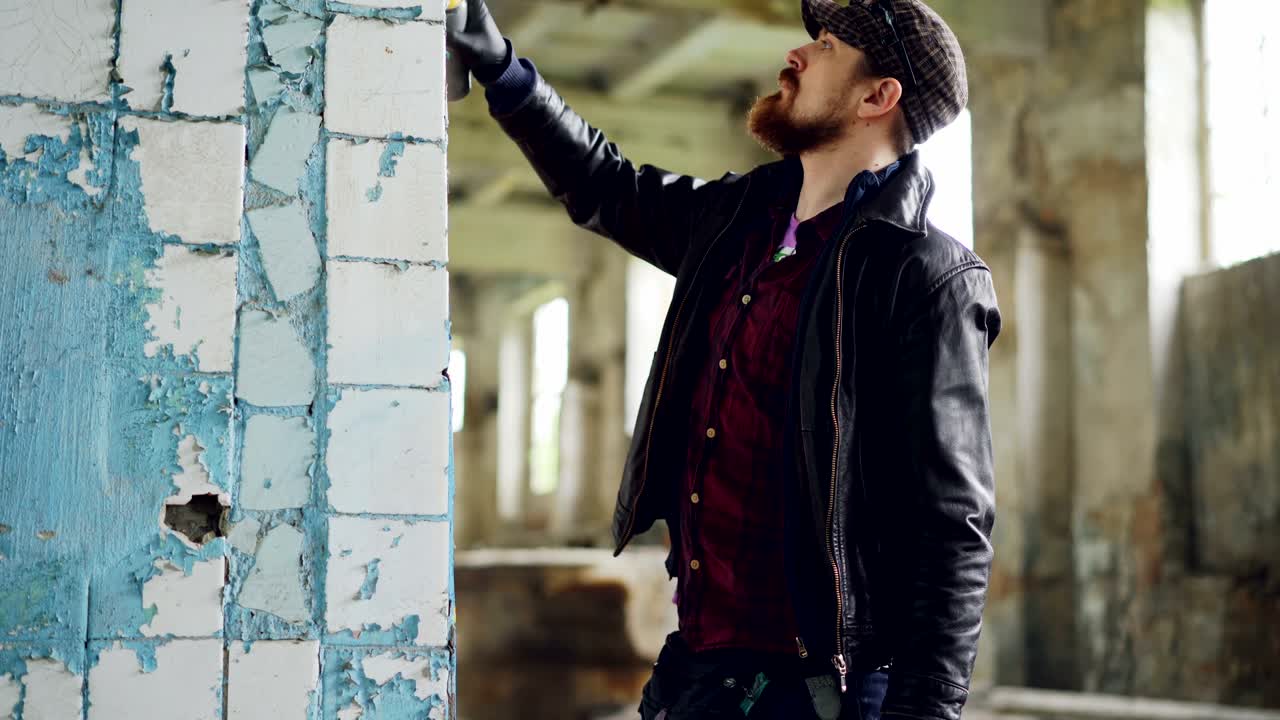 Urban street artist is standing near damaged column inside empty building and painting using aerosol paint spray. Modern art and creative young people concept.