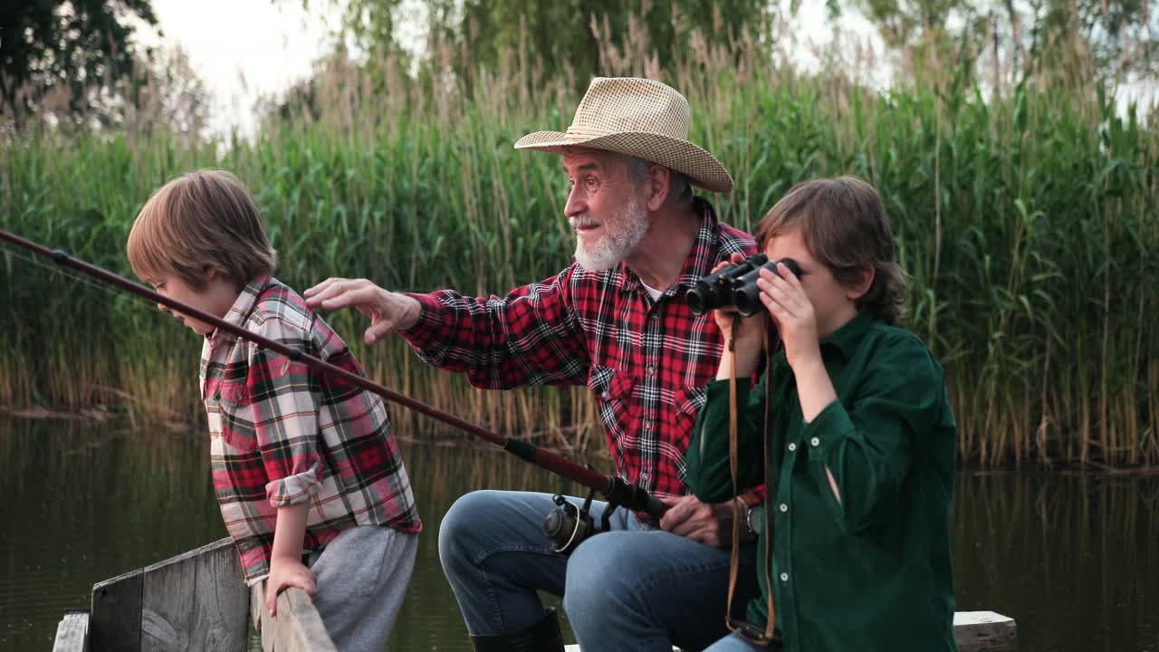 lindo adolescente usando binoculares y mostrando algo a su abuelo y a su hermano pequeño mientras pescan sentados en el muelle del lago