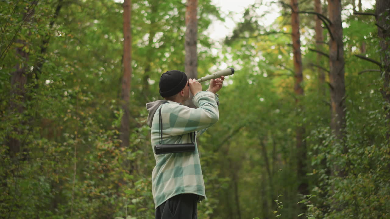 esploratore caucasico che si muove in un fitto bosco di pini, alza il binocolo per scrutare le cime degli alberi, postura attenta, zaino pieno di cinghie, ricerca mirata di uccelli, intensa luce pomeridiana, energia da tracciamento sentieri