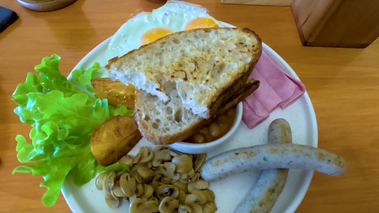 A vibrant breakfast plate with bread, sausages, beans, and greens on a wooden table in natural light