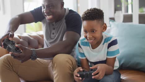 video of happy african american father and son sitting on sofa and playing video games
