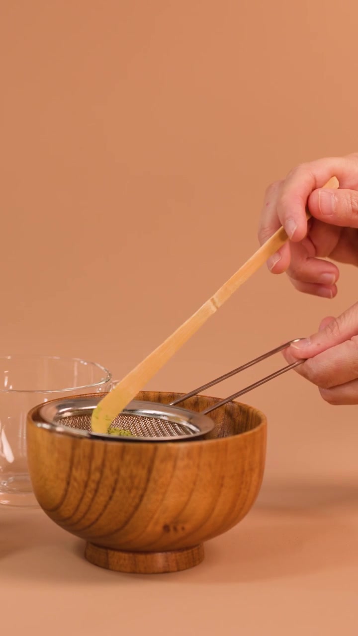 Mano setaccia vivace polvere di matcha in una ciotola di legno usando un cucchiaio di bambù e un colino a maglie fini