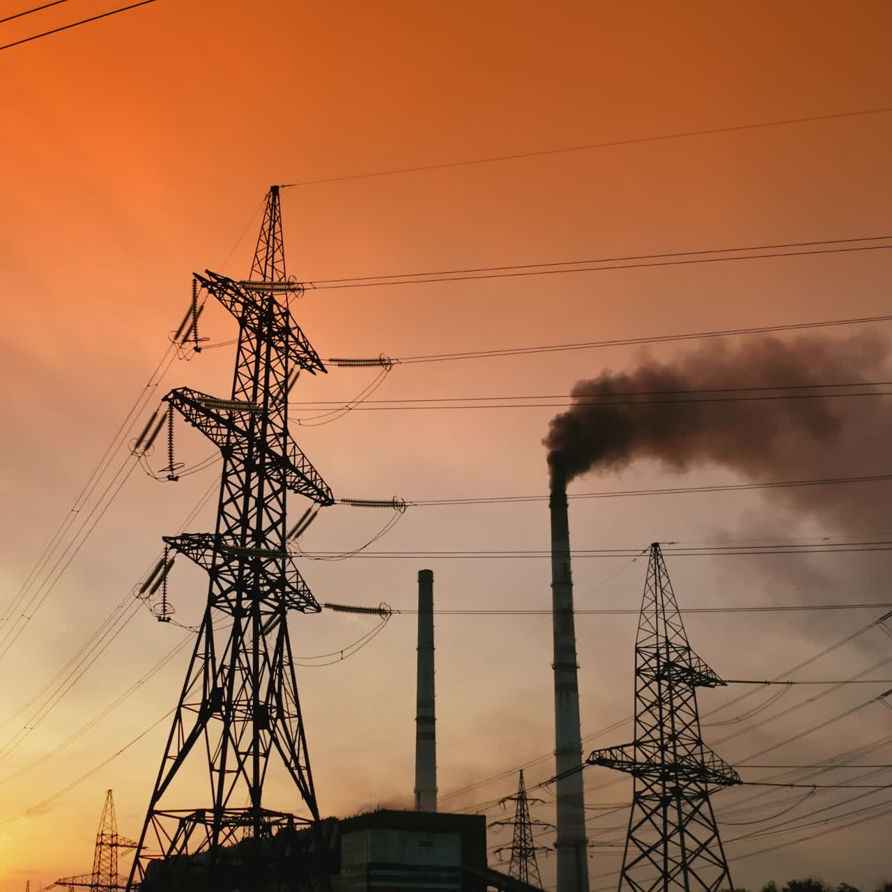 Transmission lines near smoking industrial pipe at sunset. High-voltage electric power line and dark smoke released from factory in the evening