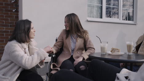 Mujer-Joven-Hablando-Con-Su-Amiga-Discapacitada-Sosteniendo-Su-Mano-En-Una-Terraza-De-Bar