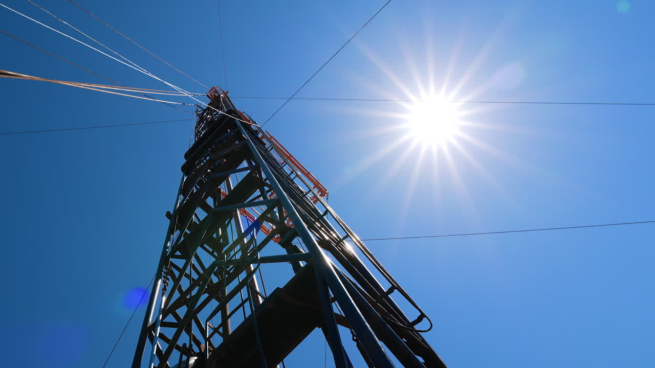 Metal derrick with equipment for oil drilling. Heavy wires stretch from the tower. Low angle view. Sun in the sky at backdrop.