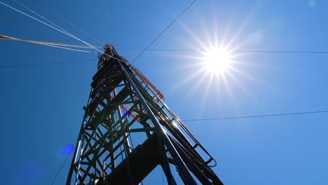 metal derrick with equipment for oil drilling. heavy wires stretch from the tower. low angle view. sun in the sky at backdrop.