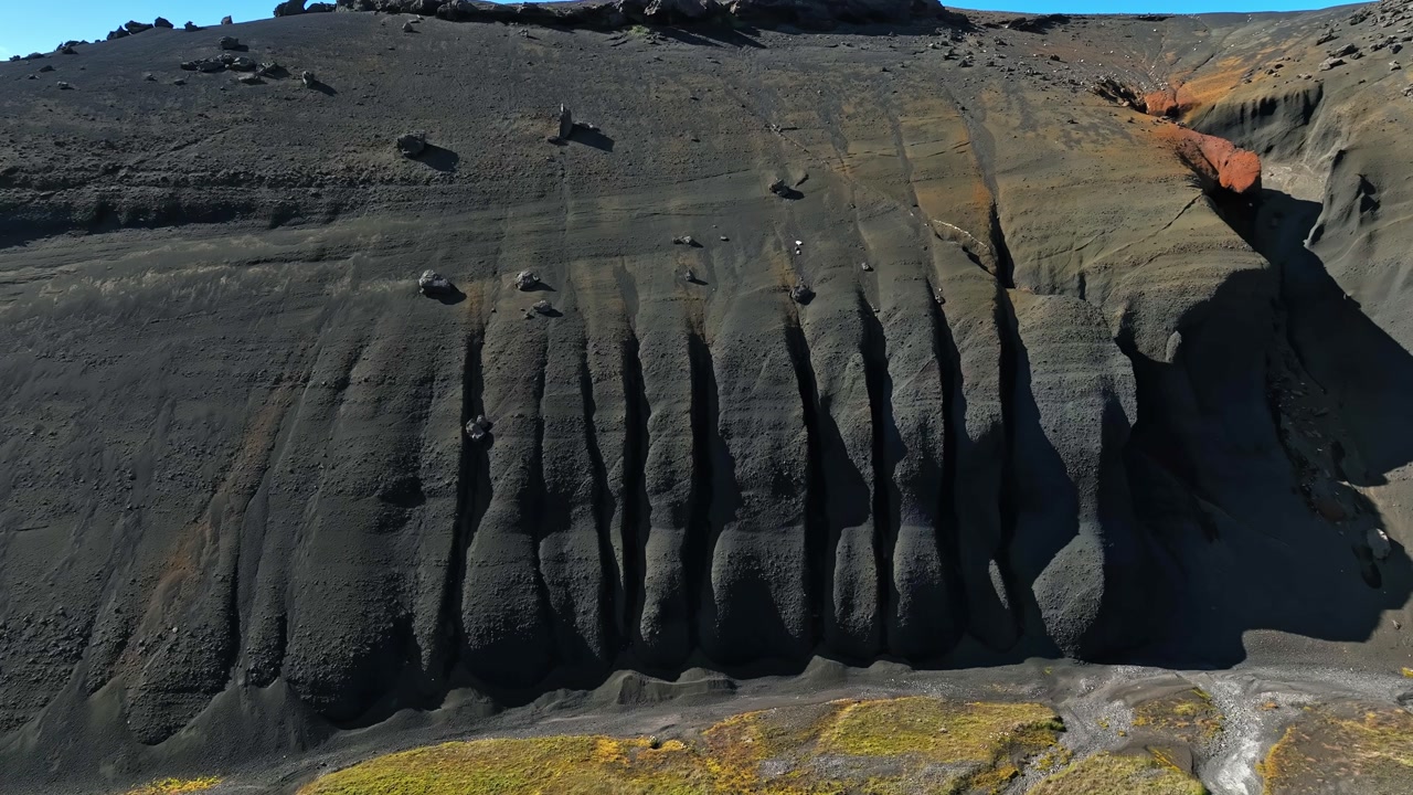 Aerial view of a black volcanic cliff carved by deep erosion grooves, with orange mineral cuts and sparse moss at the base in a stark remote geothermal landscape.