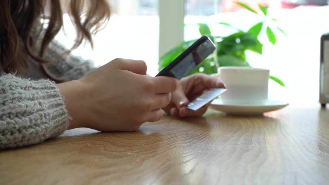 chica sosteniendo un teléfono inteligente y una tarjeta de crédito mientras está sentada en un café