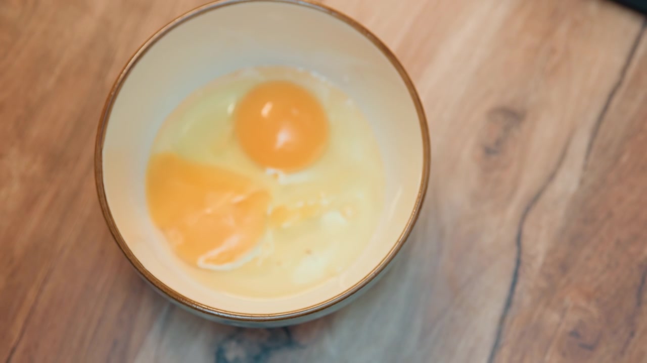 Hands cracking egg into bowl gently over ceramic mixing bowl, fingers separate shell, yolk settles into dish, home cook practicing technique, warm wooden counter, morning routine for breakfast recipe