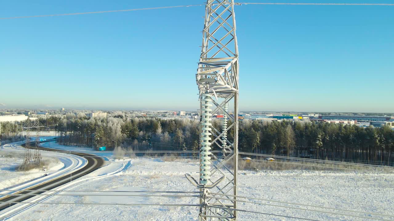 Wide aerial orbit of a frozen power line pylon reveals icy insulators above a snowy rural road, forest landscape, and distant town under clear winter sunlight