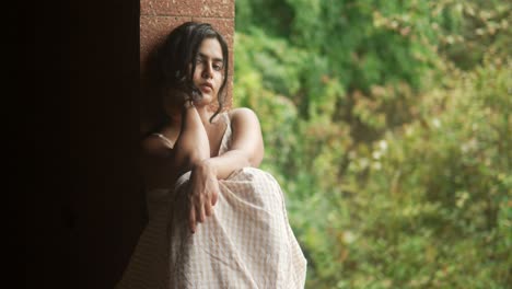 young woman in a contemplative pose by a window, looking thoughtful, with lush greenery in the background, soft focus