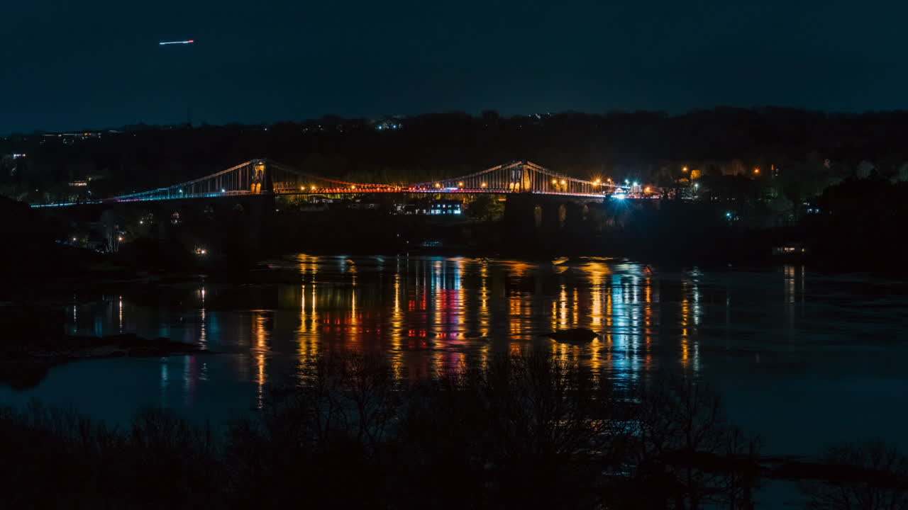 Night timelapse of Menai Bridge on Anglesey, Wales. Car light trails, storm clouds, and reflections shimmer on the water. Atmospheric and cinematic nightscape of iconic Welsh landmark