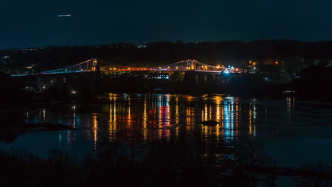 night timelapse of menai bridge on anglesey, wales. car light trails, storm clouds, and reflections shimmer on the water. atmospheric and cinematic nightscape of iconic welsh landmark