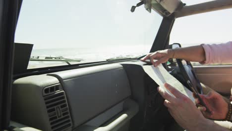 feliz pareja masculina gay caucásica sentada en el coche, leyendo el mapa y usando el teléfono inteligente en la playa
