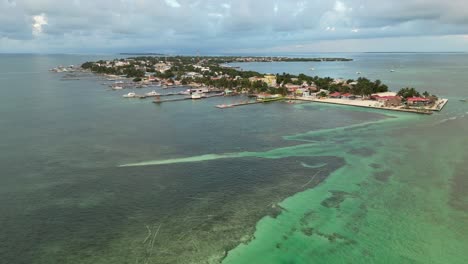 Drone-view-in-Belize-flying-over-caribbean-dark-and-light-blue-sea,-a-white-sand-caye-covered-with-palm-trees-and-restaurants-on-a-cloudy-day