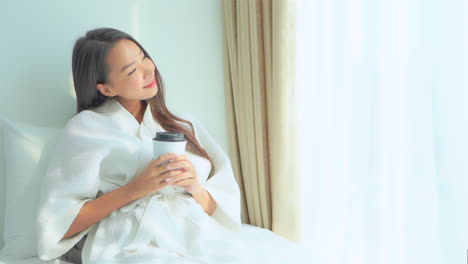 아름다운 젊은 여성이 손에 커피 한 잔을 들고 가운을 입은 호텔 스위트룸 침대에서 휴식을 취합니다.