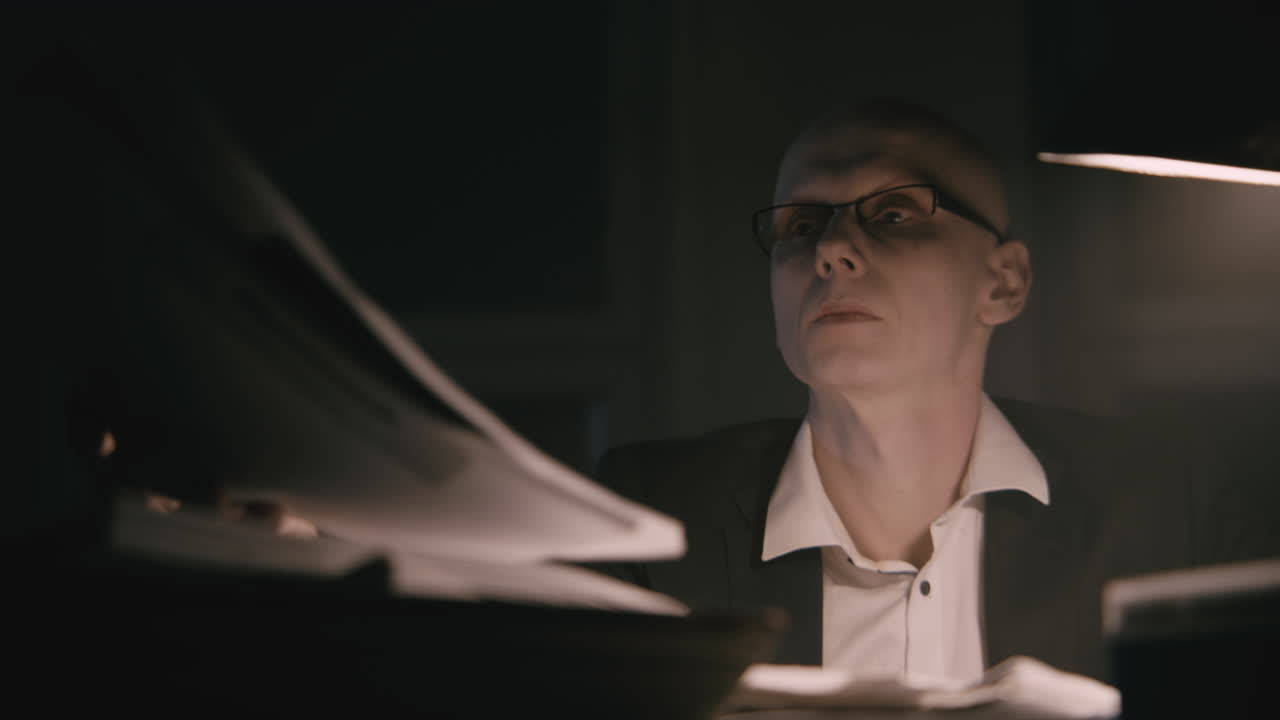 Pale bald accountant looks through the tax papers in the weird room at night