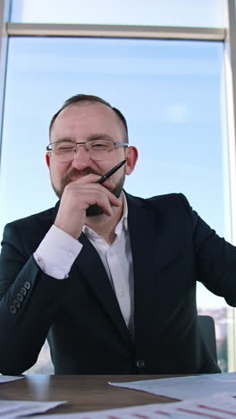 portrait of a bearded businessman at workplace. entrepreneur in glasses sitting at desk and thinking about his business. vertical video