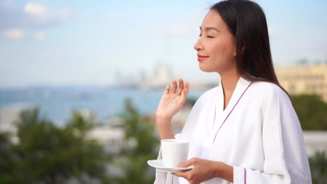 Hübsche-Junge-Asiatin-In-Weißer-Baumwollrobe-Mit-Keramiktasse-In-Der-Hand-Steht-Auf-Dem-Balkon-Mit-Blick-Auf-Die-Insel-Im-Morgenlicht