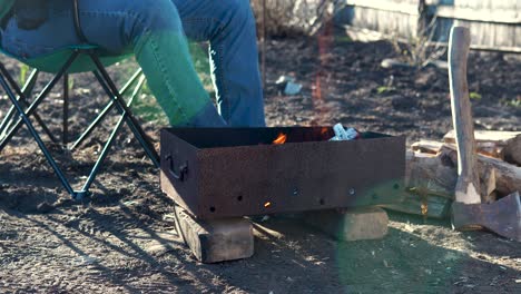 an unrecognizable man relaxedly sits on a chair in front of a bonfire in the garden. a young man is resting in the backyard, preparing a barbecue.