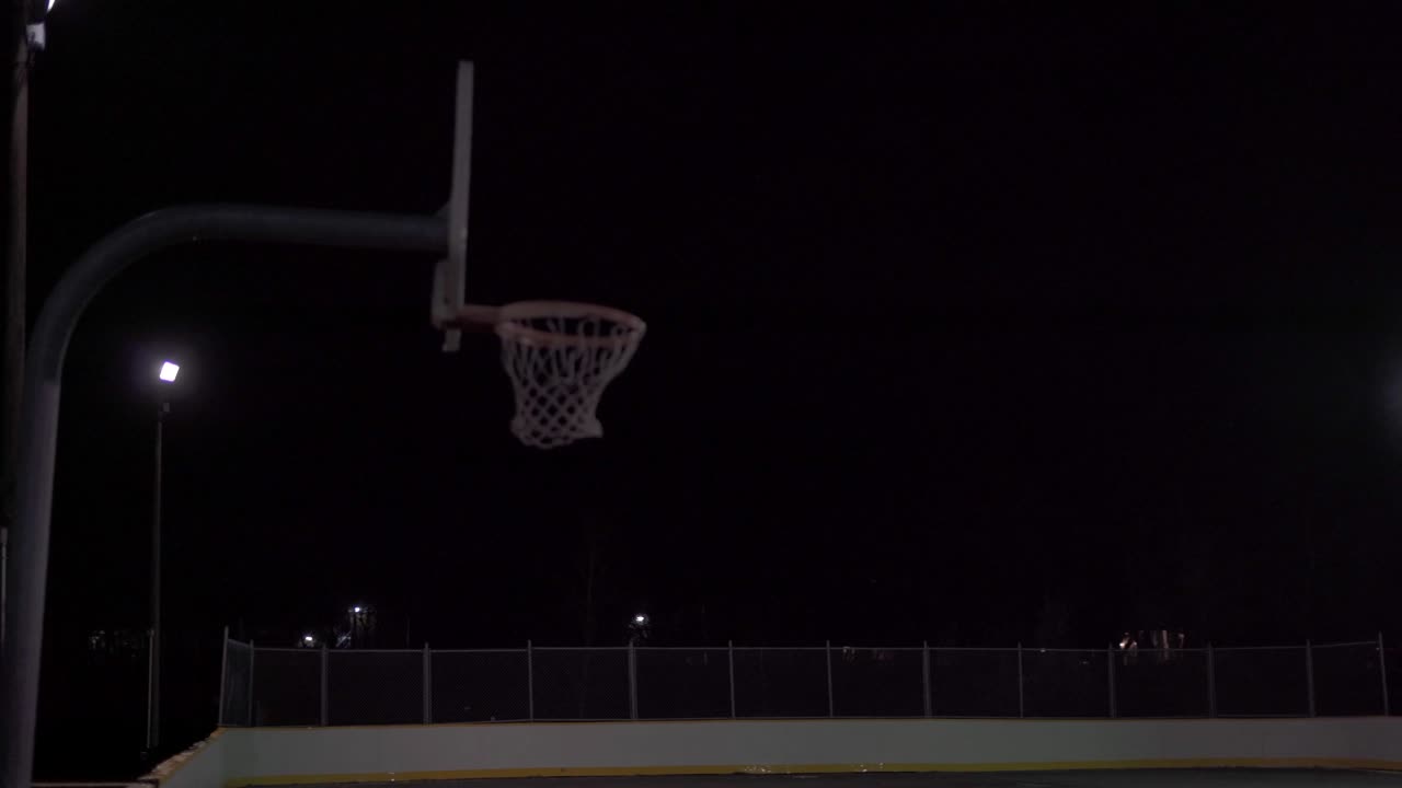 una adolescente patea el baloncesto con el pie y lo dispara en la red en una cancha oscura al aire libre con luces y lluvia