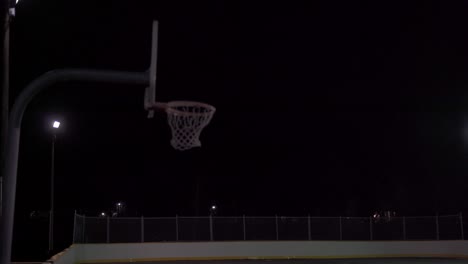 Una-Adolescente-Patea-El-Baloncesto-Con-El-Pie-Y-Lo-Dispara-En-La-Red-En-Una-Cancha-Oscura-Al-Aire-Libre-Con-Luces-Y-Lluvia