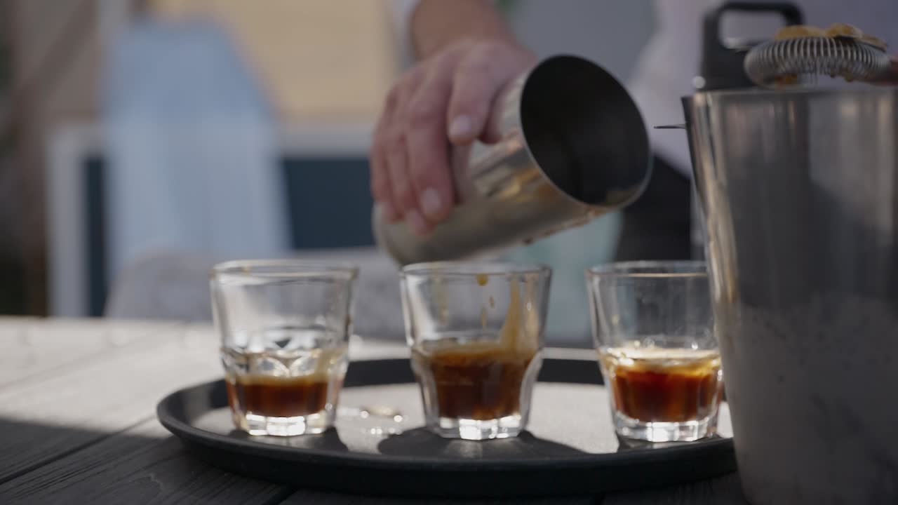 A bar tender making coffee martini cocktails and pouring it into glasses, splashing ice into them as he tries to make them even