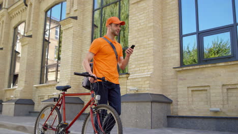 Smiling-courier-using-mobile-phone-outdoors.-Man-delivering-food-with-bike.
