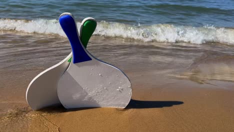 yellow little ball and beach tennis paddle rackets tilting on sandy beach with waves breaking on shore