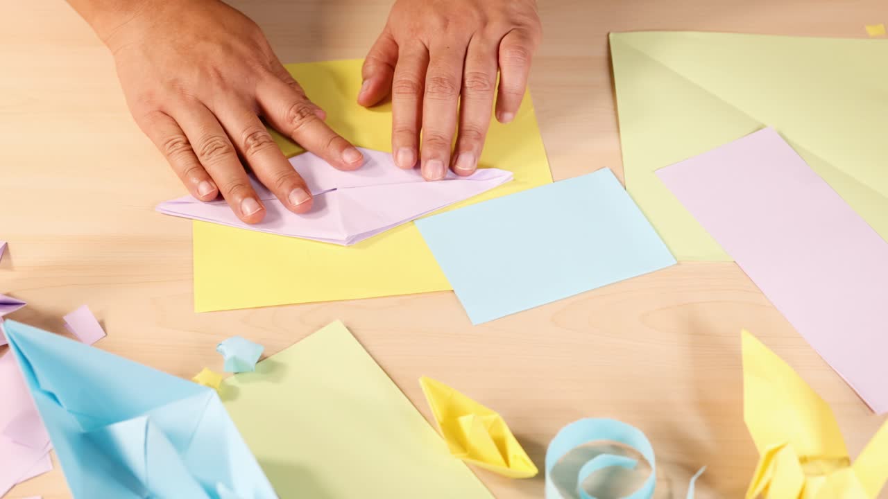 Person folds pastel origami paper on wooden table with colorful crafts, bright overhead lighting