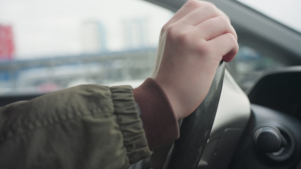 close up side view of driver hand in winter jacket lightly tapping steering wheel, calm indoor atmosphere inside parked vehicle, cityscape and natural light visible through window in soft background