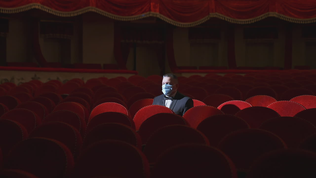 Rich man among rows of empty red chairs. Businessman in medical mask sitting in comfortable chairs alone on dark theater background. Pandemic in the world.
