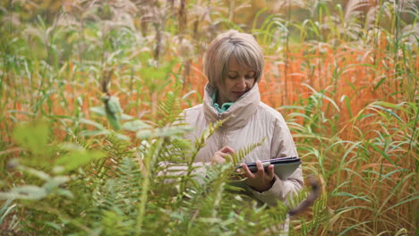 female researcher recording data on tablet while examining wild plants in autumn field, focused on scientific observations, standing amid tall grasses and ferns
