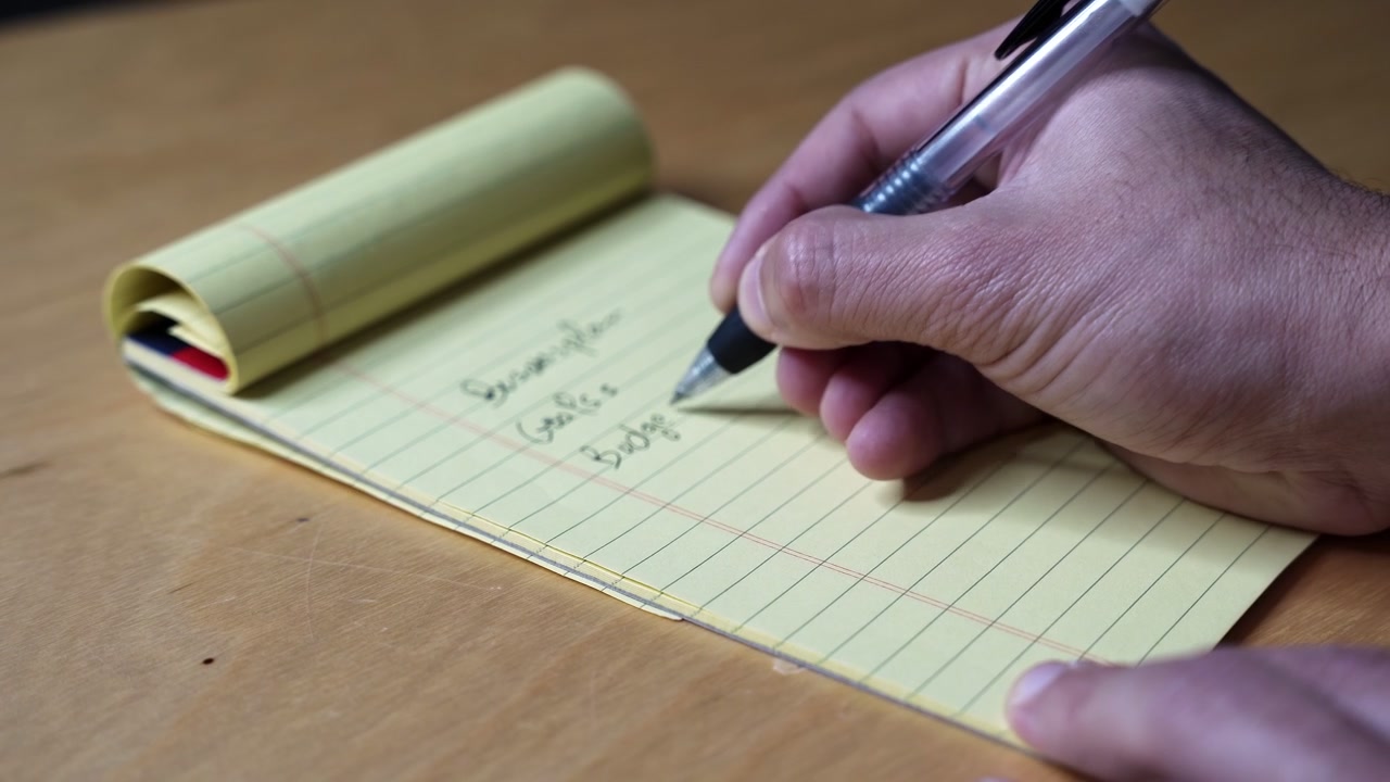 A close up shot of a person's hands writing a list of goals on a yellow legal pad. The individual uses a black ballpoint pen on a wooden desk indoors, focusing on business planning and strategy.