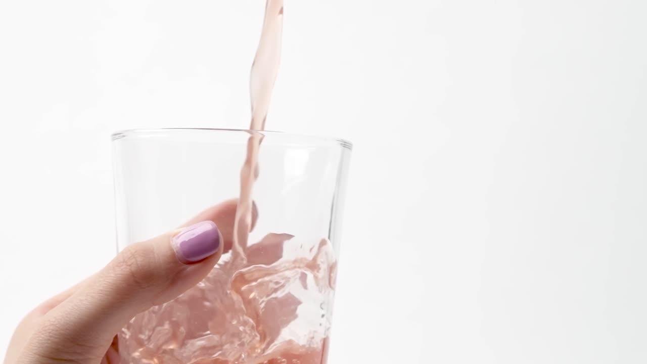 Succo rosa versato in un bicchiere vuoto tenuto da una mano femminile con smalto rosa, al rallentatore.