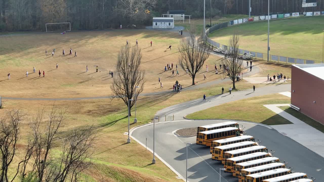 Row of parking yellow school buses in front of American school. Kids and pupils playing and doing sport exercise on soccer field of high school. Aerial wide shot