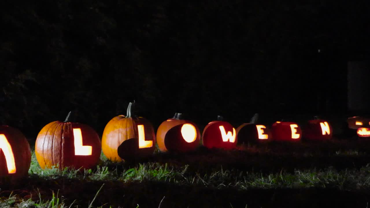 Carved out pumpkins that spell out Halloween placed in a garden during autumn night time while it's windy. Light reflects on the ground and background is dimly lit. Candles burn inside of the pumpkins