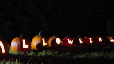 carved out pumpkins that spell out halloween placed in a garden during autumn night time while it's windy. light reflects on the ground and background is dimly lit. candles burn inside of the pumpkins