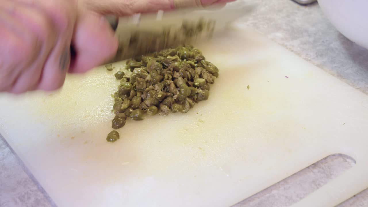 Closeup view: Woman chops capers, white chopping block with big knife