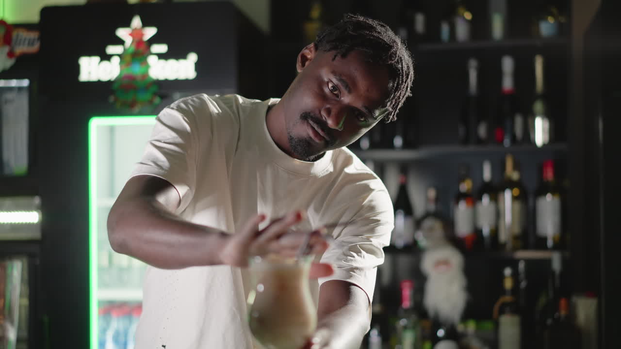 Bartender adding straw to cocktail glass, finishing drink preparation in bar. Behind, illuminated Heineken neon sign and well-stocked bar shelves, creating a lively bar atmosphere