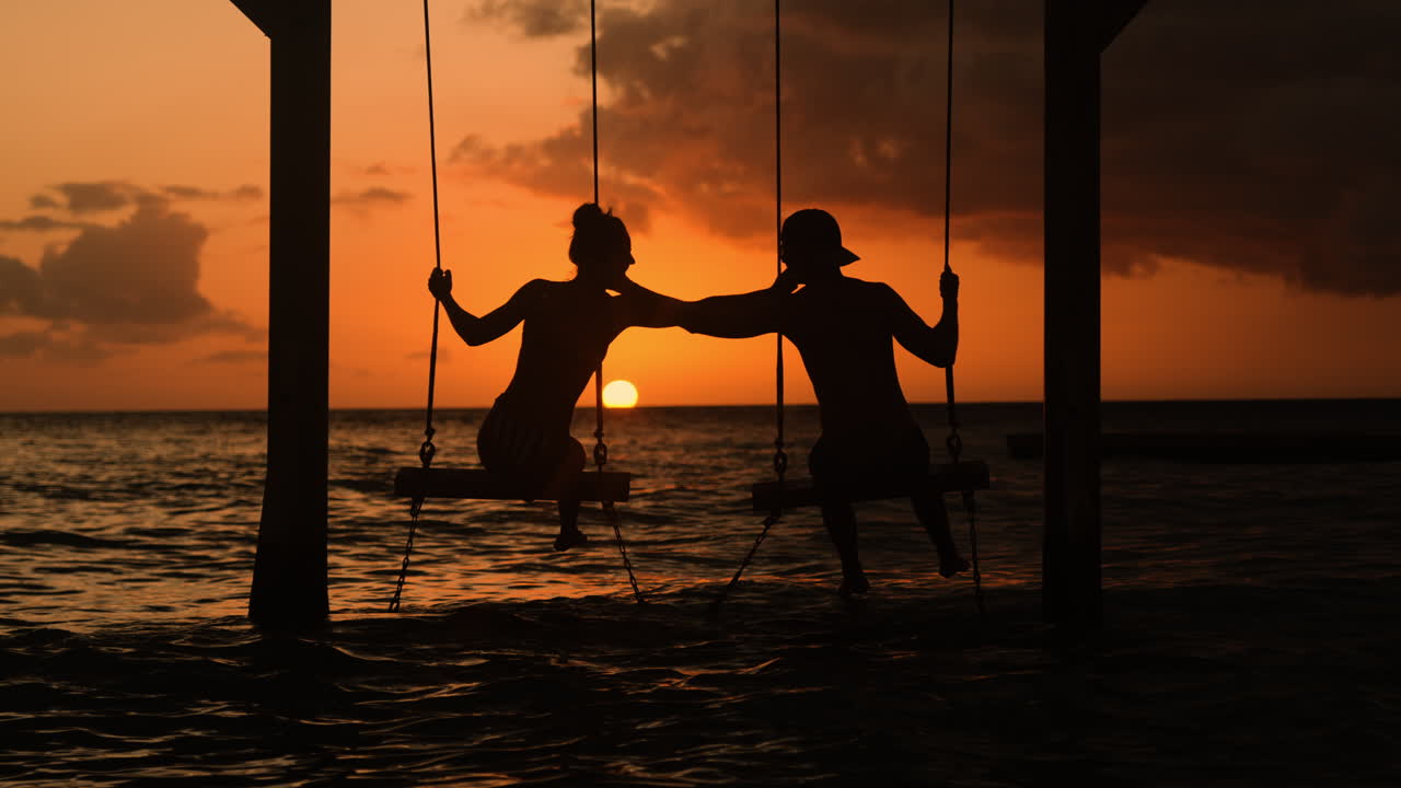 Couple Kissing on a swing while the sun sets in the ocean on the horizon, golden hour skies painting a romantic silhouette
