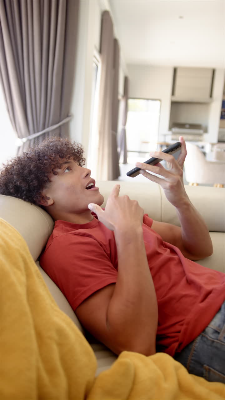 A young biracial man is reclining on a sofa, interacting with his smartphone