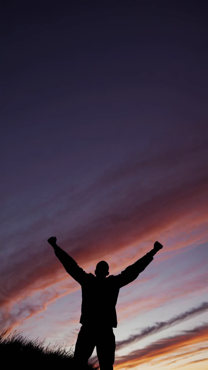 Silhouette of a person with raised arms against a vibrant sunset sky, captured from a low-angle