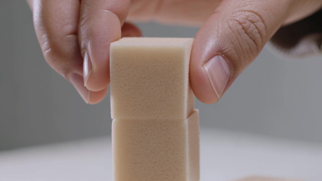 Lowering hands in dark sleeves placing held foam cube onto resting cube on white table, aligning