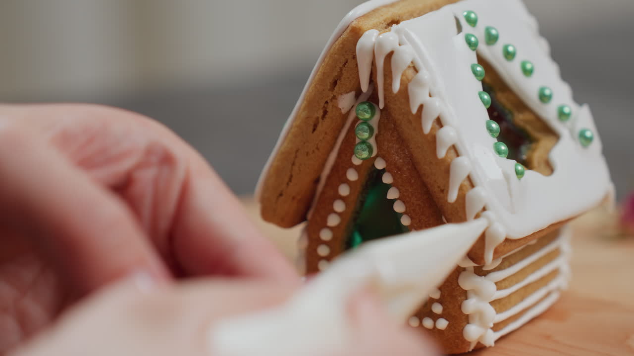 primer plano de manos que ponen helado en el techo de una casa de pan de jengibre, añadiendo detalles decorativos intrincados, estructura de galletas recién horneadas con perlas de azúcar verde en una bandeja de madera
