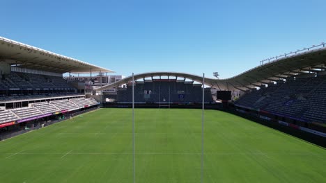 Aerial-View-of-Rugby-Stadium-Passing-Through-Goal-Posts,-Bleachers,-and-Grass-Field