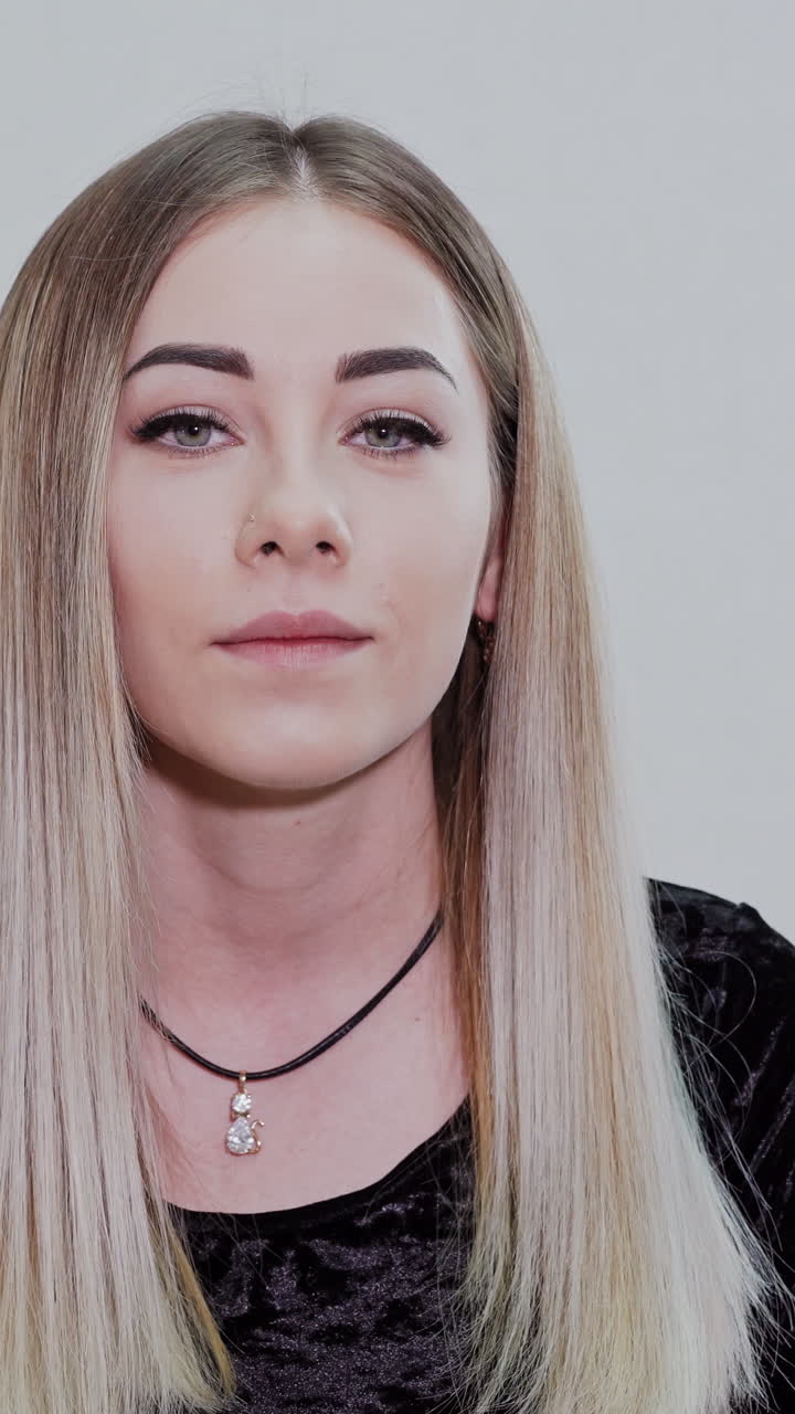 Lovely girl in studio. Portrait of a blonde girl sitting on light background and posing on camera. Natural facial features of a young woman. Vertical video