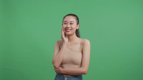 아름다운 아시아 여성이 한 손으로 얼굴을 만지고 스튜디오에서 녹색 화면 배경에서 카메라를 보고 미소 짓고 있습니다.