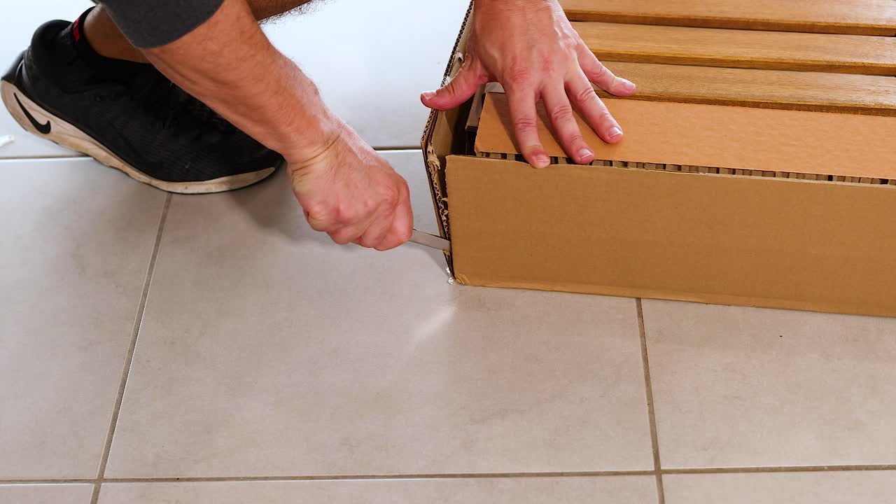 Hands carefully open a cardboard box on a tiled floor, revealing contents. Bright lighting highlights the methodical unpacking process