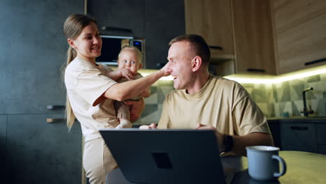 modern caucasian family at home in their kitchen. man working on laptop cheers looking at the screen. woman with baby stands behind her husband.